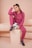 A person with braided hair is sitting on a beige pedestal, dressed in a matching pink tracksuit and black sneakers with purple laces. They are holding a soccer ball with a hand while resting their head on the other hand.