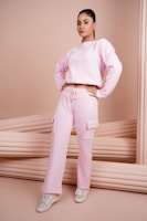 A person is standing confidently, wearing a pink sweatshirt and matching cargo pants with pockets, complemented by white sneakers. The background features a soft, neutral beige tone with cushioned elements, adding a minimalist aesthetic.