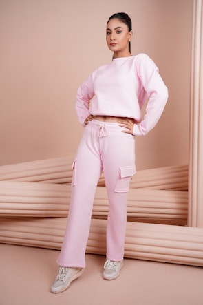 A person is standing confidently, wearing a pink sweatshirt and matching cargo pants with pockets, complemented by white sneakers. The background features a soft, neutral beige tone with cushioned elements, adding a minimalist aesthetic.