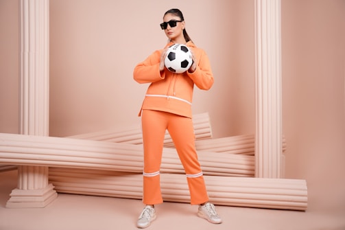 A person in an orange outfit is holding a soccer ball and wearing sunglasses, standing in front of a background adorned with architectural columns. The setting has a minimalistic design and a monochromatic color scheme.