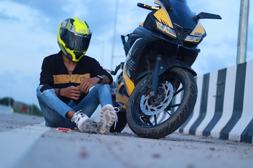 A person sits on the ground next to a motorcycle on a road with a striped barrier. The individual wears a bright yellow and black helmet, a black jacket with yellow details, ripped jeans, and white sneakers. The motorcycle is also predominantly yellow and black, matching the helmet.