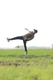 A person is performing a balancing act in an open grassy field. They are tilted sideways, with one leg lifted and both arms extended for balance. The background features a vast expanse of greenery and some distant trees, under a clear sky.