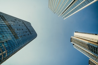 Skyscrapers appear to converge towards the center of the image as seen from below, set against a clear blue sky. The architecture is modern with extensive use of glass and steel.