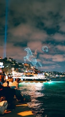 A group of people sit along the waterfront observing a light show in the sky. The night scene is illuminated by a glowing boat sailing on the water. Drones create intricate patterns of a horse and circular shapes in the sky, set against a backdrop of a cloudy night.