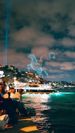 A group of people sit along the waterfront observing a light show in the sky. The night scene is illuminated by a glowing boat sailing on the water. Drones create intricate patterns of a horse and circular shapes in the sky, set against a backdrop of a cloudy night.