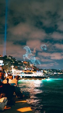 A group of people sit along the waterfront observing a light show in the sky. The night scene is illuminated by a glowing boat sailing on the water. Drones create intricate patterns of a horse and circular shapes in the sky, set against a backdrop of a cloudy night.