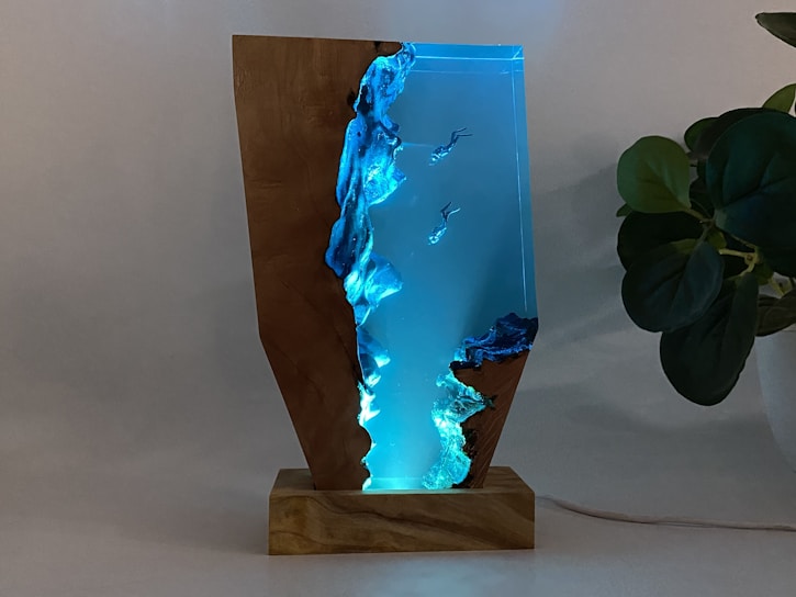 A unique decorative object made from a combination of wood and resin, illuminated with soft blue light. The resin segment mimics a watery scene with embedded figures that resemble divers or swimmers, creating an underwater effect. The natural grain and texture of the wood contrast strikingly with the translucent blue resin. Positioned on a wooden base, the piece exudes a modern and artistic vibe.