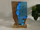 A wooden and resin sculpture on a wooden base, featuring a vivid blue resin with embedded divers, creating an underwater scene. A plant with dark green leaves is visible in the background, adding a natural element to the composition.