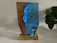 A wooden and resin sculpture on a wooden base, featuring a vivid blue resin with embedded divers, creating an underwater scene. A plant with dark green leaves is visible in the background, adding a natural element to the composition.