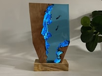 A wooden and resin sculpture on a wooden base, featuring a vivid blue resin with embedded divers, creating an underwater scene. A plant with dark green leaves is visible in the background, adding a natural element to the composition.