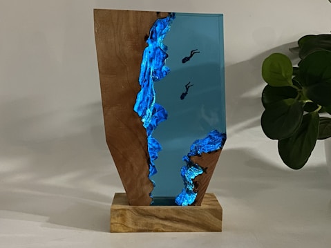 A wooden and resin sculpture on a wooden base, featuring a vivid blue resin with embedded divers, creating an underwater scene. A plant with dark green leaves is visible in the background, adding a natural element to the composition.