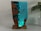 A sculptural artwork featuring a striking blend of resin and wood resembling a seascape. The piece shows translucent blue resin mimicking water with small human figures appearing to be diving or swimming, integrated into the resin. The surrounding wood adds an organic texture, enhancing the visual contrast. It is positioned on a solid wood base and placed next to a leafy green plant in a white pot, set against a neutral background.
