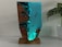 A sculptural artwork featuring a striking blend of resin and wood resembling a seascape. The piece shows translucent blue resin mimicking water with small human figures appearing to be diving or swimming, integrated into the resin. The surrounding wood adds an organic texture, enhancing the visual contrast. It is positioned on a solid wood base and placed next to a leafy green plant in a white pot, set against a neutral background.