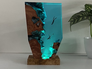A sculptural artwork featuring a striking blend of resin and wood resembling a seascape. The piece shows translucent blue resin mimicking water with small human figures appearing to be diving or swimming, integrated into the resin. The surrounding wood adds an organic texture, enhancing the visual contrast. It is positioned on a solid wood base and placed next to a leafy green plant in a white pot, set against a neutral background.