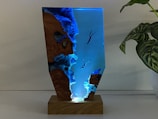 A sculpture featuring a vertical, resin-like structure mounted on a wooden base. The piece has a transparent, blue-tinted section with embedded figures of divers swimming towards the bottom. The sides feature textured, rocky formations resembling a cliff or cave. To the right of the sculpture, there is a green plant with variegated leaves.