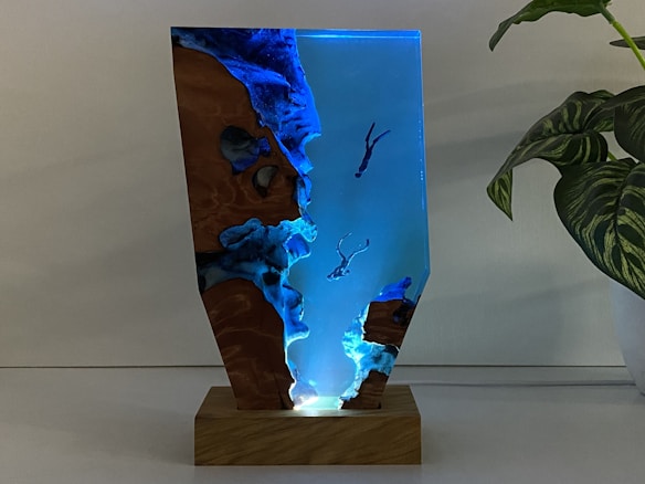 A sculpture featuring a vertical, resin-like structure mounted on a wooden base. The piece has a transparent, blue-tinted section with embedded figures of divers swimming towards the bottom. The sides feature textured, rocky formations resembling a cliff or cave. To the right of the sculpture, there is a green plant with variegated leaves.