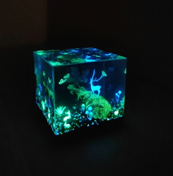 A luminescent cube with an enchanting scene inside, featuring a glowing blue deer standing on a mossy log surrounded by small plants and abstract shapes. The scene is vibrant, with various shades of blue and green illuminating in the dark.