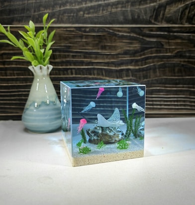 A cube-shaped object resembling an aquarium contains miniature sea life models with small stingrays and colorful jellyfish. Notably, the bottom has sandy textures, and the interior is dotted with tiny green plants and rocks. In the background, a vase with fresh green leaves stands, set against a wavy wooden pattern wall.