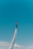A person is elevated high in the sky using a water jetpack, surrounded by clear blue skies and a few clouds below. The water jet creates a powerful vertical spray.