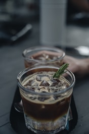 Two glasses filled with iced coffee are placed on a dark surface, with one glass having a sprig of rosemary as a garnish. The beverage is a mixture of cream and dark coffee, creating a marbled effect. The background is blurred, focusing attention on the drinks.