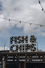 A vintage-style sign advertising 'Fish & Chips' is prominently displayed against a cloudy sky. The sign is decorated with light bulbs, giving it a retro look. Below the sign, there is a menu board listing various seafood options with prices. Colorful string lights hang above, adding a festive touch.