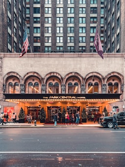 A grand hotel entrance with an illuminated marquee displaying 'The Park Central Hotel'. The building's architecture includes tall windows and decorative arches. Two flags, including an American flag, are displayed above the entrance. People are gathered in front of the building, and Christmas trees with lights are positioned by the entrance. A black SUV is parked on the street in front.
