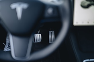 A close-up view of a car's steering wheel with a recognizable logo, foot pedals, and part of a dashboard display. The steering wheel is in the foreground, while two metallic pedals for acceleration and braking are visible beneath. A portion of a touchscreen is visible on the right side.