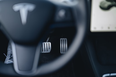 A close-up view of a car's steering wheel with a recognizable logo, foot pedals, and part of a dashboard display. The steering wheel is in the foreground, while two metallic pedals for acceleration and braking are visible beneath. A portion of a touchscreen is visible on the right side.