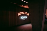 Neon signs illuminate a dark interior space, with the words 'Vienna House Mokotow Warsaw' displayed in glowing letters. The lighting casts a moody, atmospheric feel against the ribbed wall behind. A large, angular structural column partially frames the scene on the right.