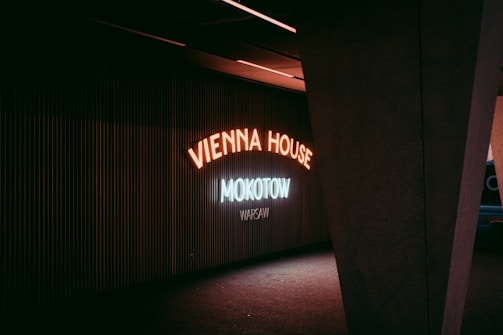 Neon signs illuminate a dark interior space, with the words 'Vienna House Mokotow Warsaw' displayed in glowing letters. The lighting casts a moody, atmospheric feel against the ribbed wall behind. A large, angular structural column partially frames the scene on the right.