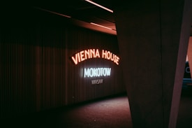Neon signs illuminate a dark interior space, with the words 'Vienna House Mokotow Warsaw' displayed in glowing letters. The lighting casts a moody, atmospheric feel against the ribbed wall behind. A large, angular structural column partially frames the scene on the right.