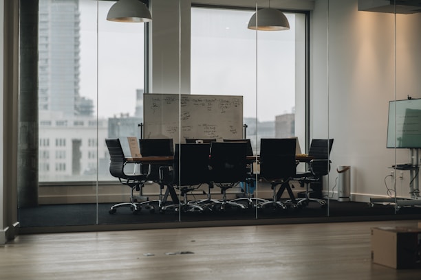 A modern conference room with a long table and several black office chairs. The room has glass walls and windows revealing an urban exterior. A whiteboard with handwritten notes stands at the end of the table. The atmosphere is structured with neutral tones.