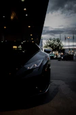 Side profile of a black luxury car featuring tinted windows reflecting a cityscape at dusk with red highlights.