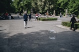 A large bubble is floating in the air in an open park area. Several people, including a man in a blue jacket and a hat, are walking along the paved path surrounded by green bushes and tree-lined walkways. Some are sitting and standing in the distance, enjoying the outdoor setting.