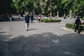 A large bubble is floating in the air in an open park area. Several people, including a man in a blue jacket and a hat, are walking along the paved path surrounded by green bushes and tree-lined walkways. Some are sitting and standing in the distance, enjoying the outdoor setting.
