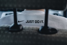 A close-up view of the back of a person's white shirt, featuring a prominent black Nike logo and the slogan 'JUST DO IT.' The image is taken from behind a car seat, suggesting the person is sitting inside a vehicle. The overall focus is on the branding and slogan.