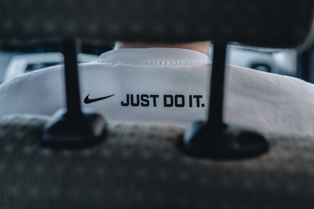 A close-up view of the back of a person's white shirt, featuring a prominent black Nike logo and the slogan 'JUST DO IT.' The image is taken from behind a car seat, suggesting the person is sitting inside a vehicle. The overall focus is on the branding and slogan.