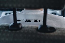 A close-up view of the back of a person's white shirt, featuring a prominent black Nike logo and the slogan 'JUST DO IT.' The image is taken from behind a car seat, suggesting the person is sitting inside a vehicle. The overall focus is on the branding and slogan.