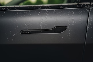 Close-up of a sleek car door handle with water droplets reflecting electric blue highlights on a dark carbon fiber background