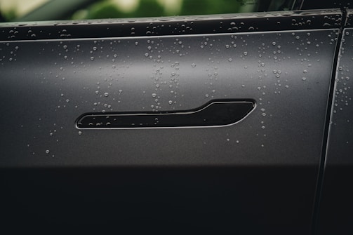 Close-up of a sleek car door handle with water droplets reflecting electric blue highlights on a dark carbon fiber background