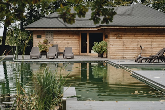 A rustic wooden house is situated beside a natural swimming pool. The house is surrounded by lush greenery, and several lounge chairs are arranged on a wooden deck. Potted plants add to the natural ambiance, and the water reflects the house and surrounding trees.