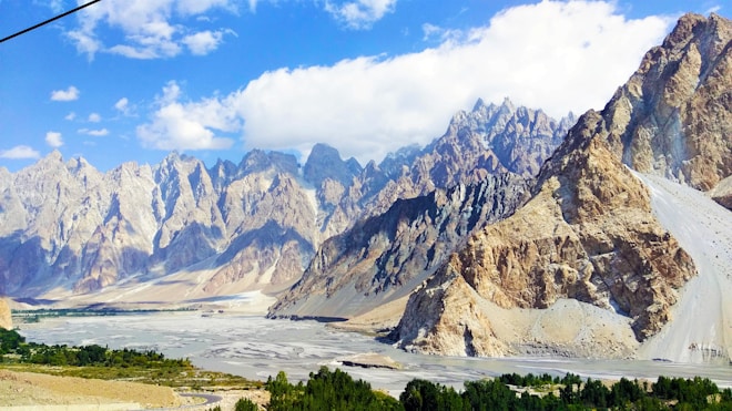 A breathtaking view of rugged mountains with jagged peaks, contrasting against a bright blue sky. Sparse clouds float above, adding depth to the scenery. At the base, a wide riverbed seemingly parched, surrounded by patches of greenery adds a hint of life to the arid landscape.