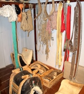 A collection of traditional harness equipment, including ropes, harness pads, and leather straps, hangs on a wall. There are several coils of rope and various colored straps. Wooden and leather saddles are placed on the floor, alongside wooden boxes with tools and accessories.