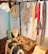 A collection of traditional harness equipment, including ropes, harness pads, and leather straps, hangs on a wall. There are several coils of rope and various colored straps. Wooden and leather saddles are placed on the floor, alongside wooden boxes with tools and accessories.