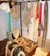 A collection of traditional harness equipment, including ropes, harness pads, and leather straps, hangs on a wall. There are several coils of rope and various colored straps. Wooden and leather saddles are placed on the floor, alongside wooden boxes with tools and accessories.