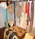 A collection of traditional harness equipment, including ropes, harness pads, and leather straps, hangs on a wall. There are several coils of rope and various colored straps. Wooden and leather saddles are placed on the floor, alongside wooden boxes with tools and accessories.