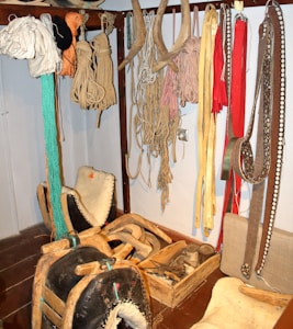 A collection of traditional harness equipment, including ropes, harness pads, and leather straps, hangs on a wall. There are several coils of rope and various colored straps. Wooden and leather saddles are placed on the floor, alongside wooden boxes with tools and accessories.
