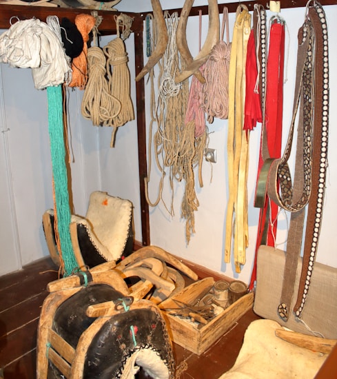 A collection of traditional harness equipment, including ropes, harness pads, and leather straps, hangs on a wall. There are several coils of rope and various colored straps. Wooden and leather saddles are placed on the floor, alongside wooden boxes with tools and accessories.