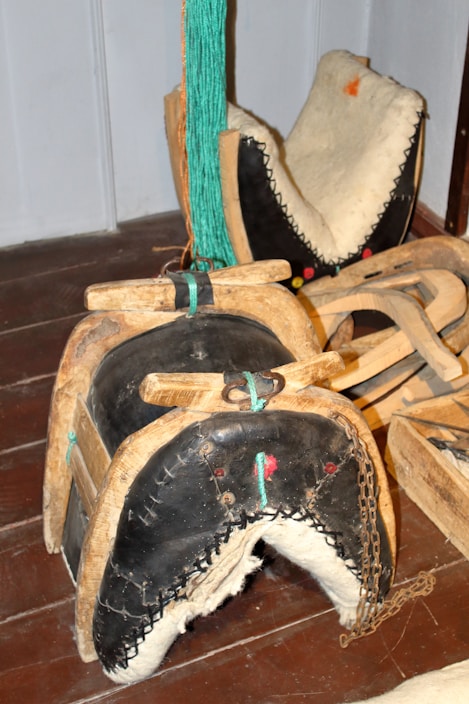 An intricately crafted wooden and leather horse saddle with a rustic appearance. The saddle features black leather padding with white stitching and is accented with turquoise rope and metal hardware. It is surrounded by other similar wooden saddle frames, resting on a dark wooden floor.