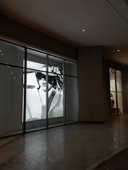 A black and white photograph of a person wearing sunglasses, displayed prominently in a large shop window. The image is highly stylized and captures an element of fashion and mystery.
